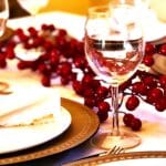 A festive holiday table setting featuring a clear wine glass in focus. The table is decorated with red berry garlands, golden charger plates, white dinner plates, and neatly folded white napkins tied with twine for a rustic touch. The scene evokes a warm and elegant Christmas dining atmosphere.