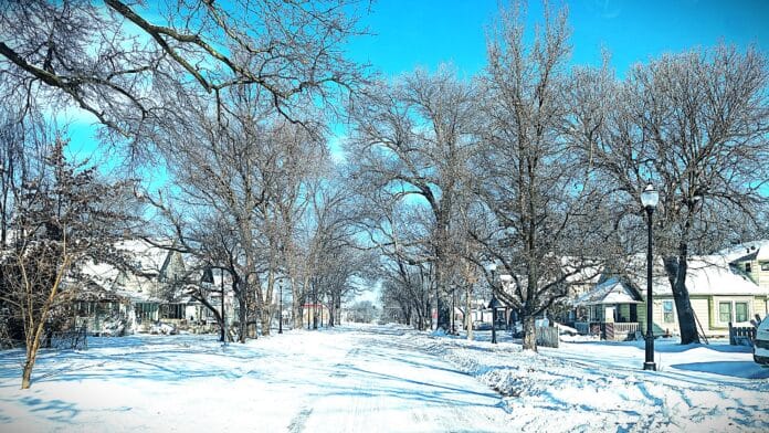 A residential street in Topeka covered in snow, with tree branches arching over the road. Houses line both sides of the street, and a clear blue sky provides a bright backdrop. Snow piles up along the edges, and the road shows signs of light plowing, with patches of snow remaining