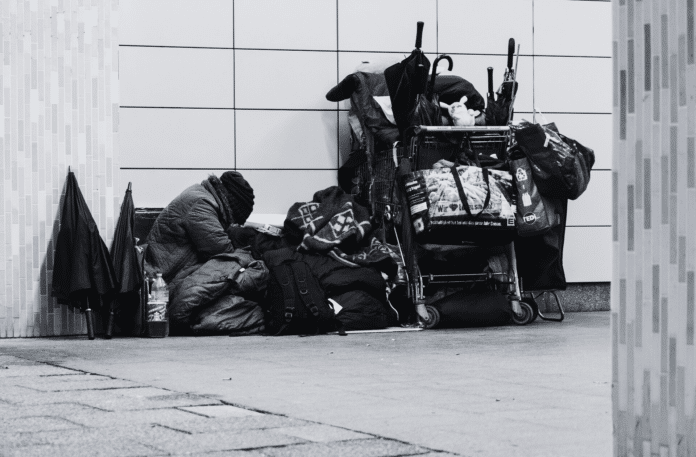 A homeless individual sits on the ground beside their belongings, including a shopping cart filled with blankets, bags, and personal items, symbolizing the challenges of homelessness in urban areas.