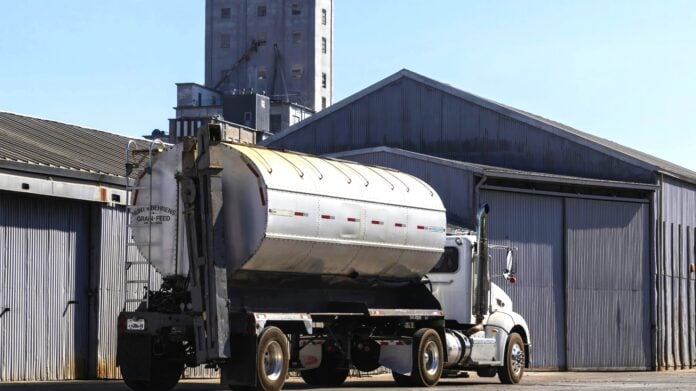 pexels-robertkso-18468412 A white semi-truck with a silver tanker trailer labeled "Hunt & Behrens Grain + Feed" is parked in front of a large corrugated metal warehouse under a clear blue sky, with a grain elevator visible in the background.